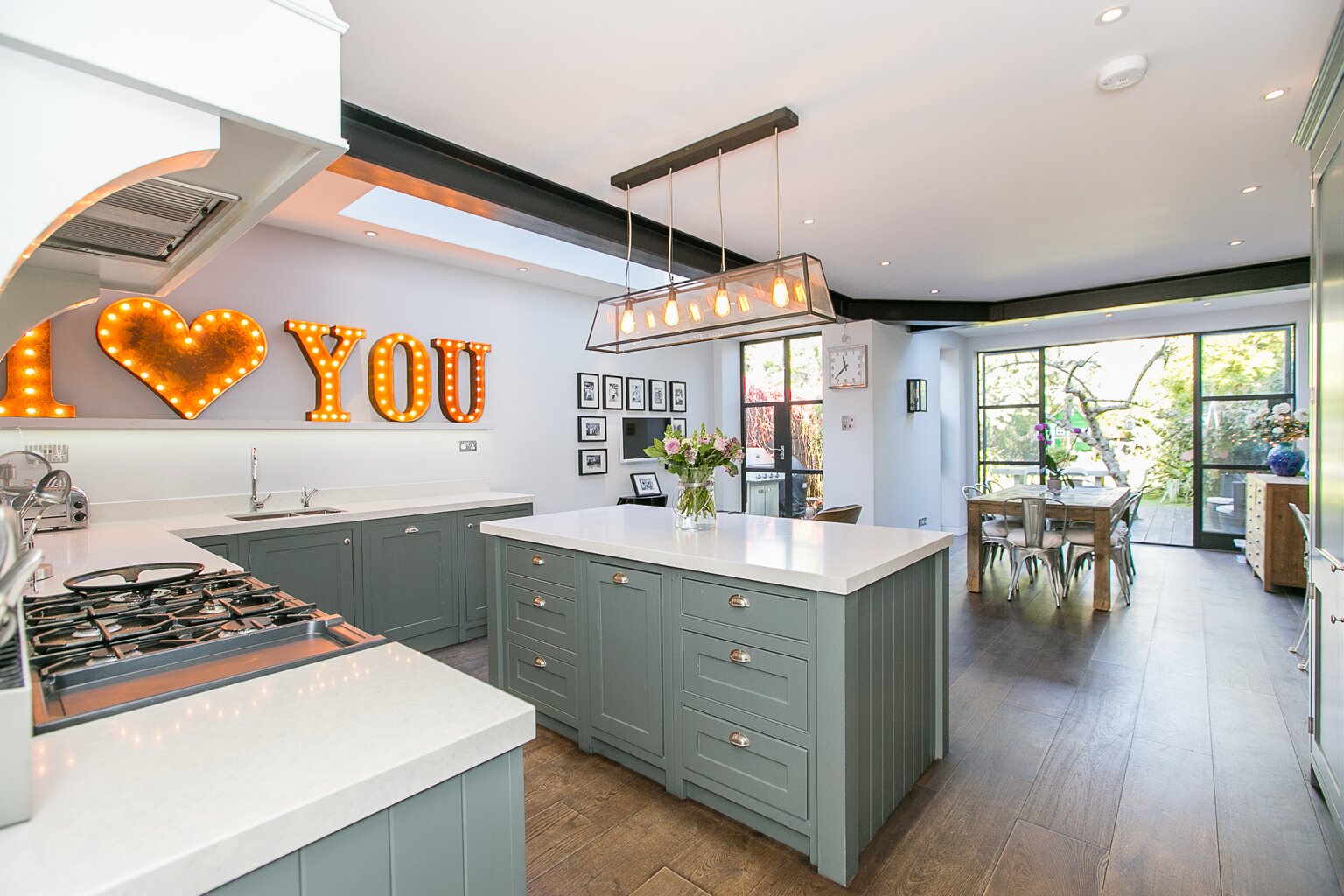 Grey-green fitted kitchen and island beside an open dining area and black-framed glazed doors in Brondesbury, London