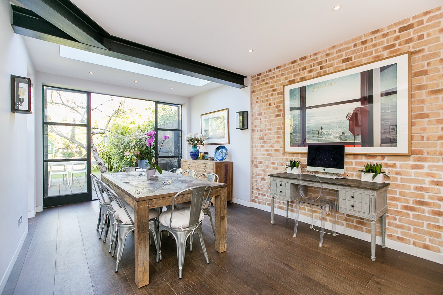 Dining area in Brondesbury, London with a brick accent wall, dark floor, rooflight and large black-framed glazed doors